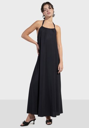 Robe maxi noire avec décolleté halter, corps froncé, tissu fluide et texture lisse. Portée avec des sandales à talons noires décorées de nœuds.