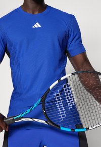 Blaues sportliches T-Shirt mit gerippter Textur, versehen mit einem weißen Logo, kombiniert mit einem Tennisschläger. Kurze Ärmel, eng anliegende Passform.