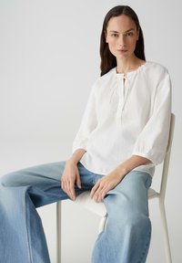 Blusa bianca in lino con scollo rotondo, maniche corte a palloncino e dettaglio cravatta al collo, abbinata a jeans a gamba larga di un leggero blu.