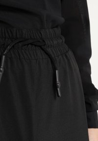 Svarta träningbyxor med en slät textur, med resårmidja och dragsko. Utmärkande svarta snoddar i ändarna.