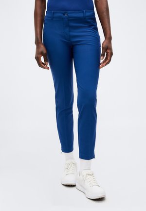 Personne portant un pantalon bleu slim, des chaussettes blanches et des baskets blanches, debout sur un fond clair uni.