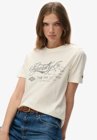 Ung kvinde med lyst brunt hår iført en cremefarvet t-shirt med logo og blå jeans med et brunt vævet bælte, poserer foran en ensfarvet hvid baggrund.