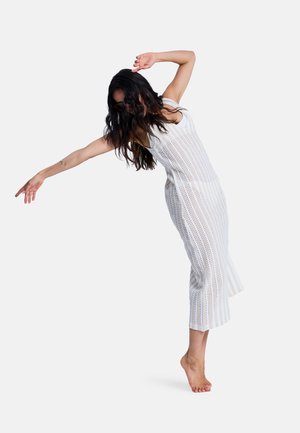 Weißes Strickkleid mit vertikalen Streifen, ärmelloses Design, verfügt über Ausschnittmuster und eine Midi-Länge, getragen barfuß von einem Modell in einer Pose.