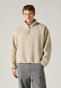 Μπεζ φλις μπλούζα με υψηλό γιακά και κλείσιμο με μισό φερμουάρ, με υφή στην επιφάνεια και ευθεία ρίγα στο κάτω μέρος. Συνδυάζεται με γκρι παντελόνια cargo.