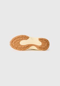Suela de zapatilla en goma crema y marrón claro, con un patrón texturizado que incluye surcos y acentos circulares para ofrecer tracción.