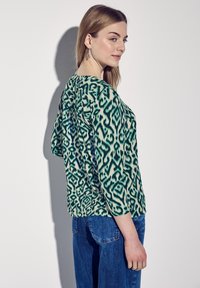 Haut vert et crème à motifs avec des manches trois-quarts, assorti à un jean en denim bleu. Le haut présente un design abstrait et une texture douce.