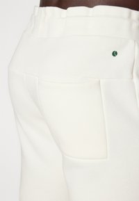 Weiße Sweatpants aus Baumwollmischung mit einem gerippten elastischen Bund, ausgestattet mit einer einzelnen Gesäßtasche und einem kleinen grünen Logo-Akzent.