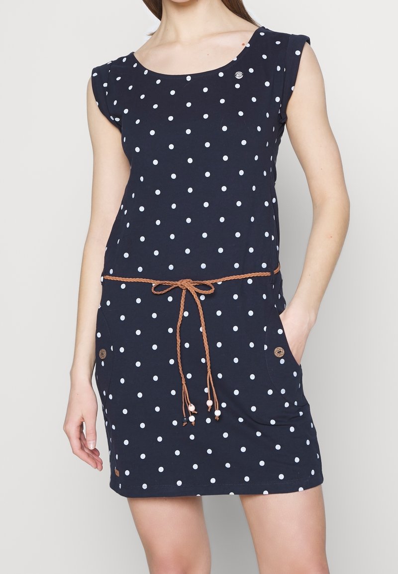 Vrouw draagt een marineblauwe mouwloze jurk met witte polka dots, een bruine gevlochten riem gestrikt in de taille en zakken met knoopdetails.