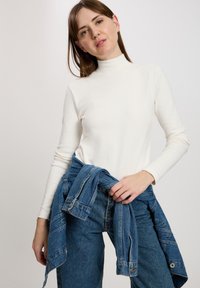Kremowy sweter z golfem i długim rękawem, zestawiony z niebieską kurtką dżinsową związaną w talii, z detalami w postaci guzików i kieszeni.