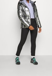 Personne portant une doudoune métallisée argentée, un haut lavande à fermeture éclair au quart, des leggings noirs, des chaussettes blanches et des baskets noires avec des lacets turquoise.