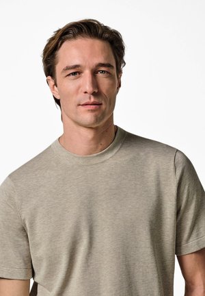 Uomo adulto con capelli castano chiaro che indossa una maglietta beige a maniche corte e collo tondo, guardando direttamente avanti su uno sfondo bianco.