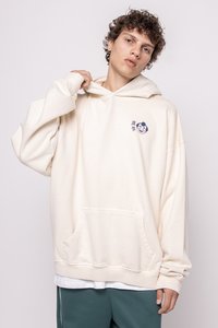 Sudadera de corte oversized en color crema, hecha de una tela suave, que presenta un bolsillo frontal y un gráfico de un personaje sonriente en la parte izquierda del pecho.