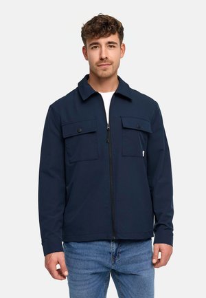 Młody mężczyzna z krótkimi kręconymi włosami, ubrany w granatową kurtkę z frontowym zamkiem błyskawicznym i zapinanymi na guziki kieszeniami na piersi, w połączeniu z niebieskimi dżinsami, stojący na białym tle.