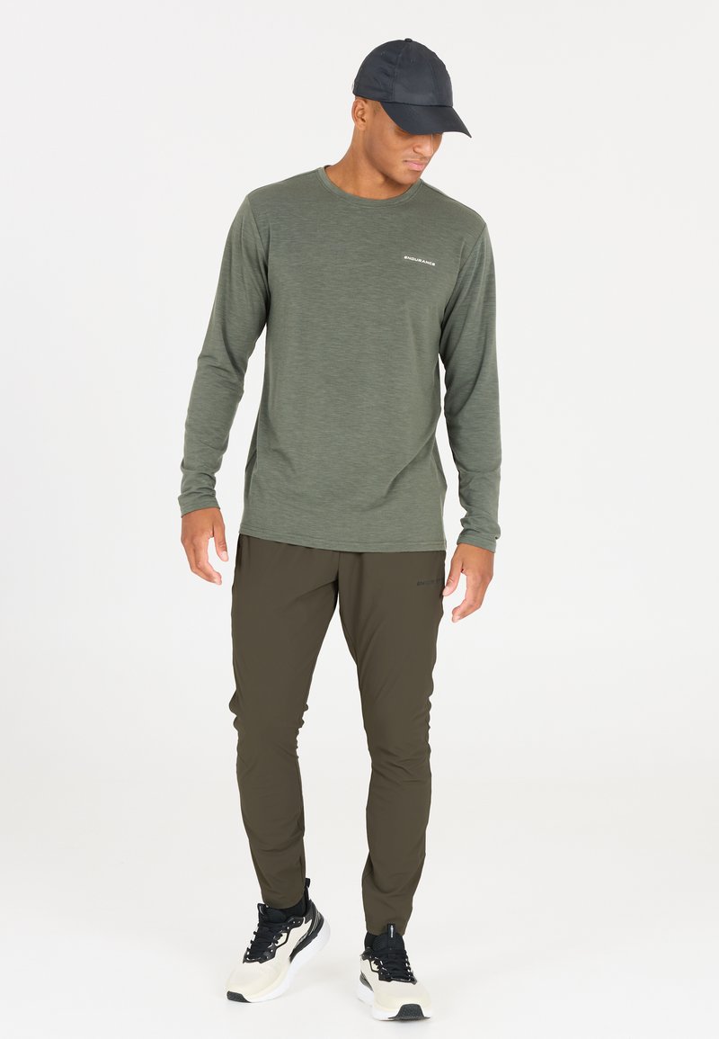 Langsleeve, olijfgroen shirt met een ronde hals en een vaag logo. Gecombineerd met slim-fit, donker olijfkleurige broek en zwarte sportschoenen.