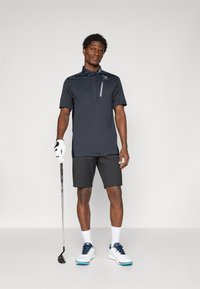 Sötétkék pólóing rövid ujjakkal, cipzáras részlettel és logóval, fekete rövidnadrággal és fehér golfsapkában. Golf felszereléssel áll.