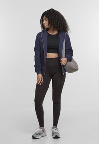 Felpa con zip blu navy, top corto nero, leggings marroni a vita alta, borsa trapuntata grigia e scarpe da ginnastica grigie con dettagli bianchi.