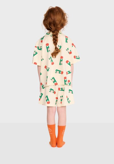 Enfant aux longs cheveux roux tressés porte une chemise crème et un short avec des motifs verts et rouges, assortis à des chaussettes orange vif, debout sur fond blanc.
