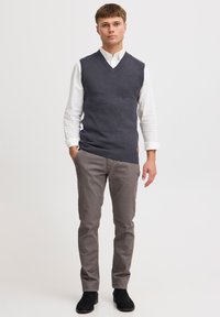 Graue Strickweste mit V-Ausschnitt, getragen über einem weißen Langarmshirt. Kombiniert mit grauen Hosen und schwarzen Schuhen. Saubere, moderne Silhouette.