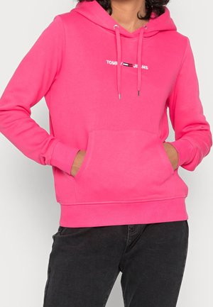 Person wearing bright pink Tommy Jeans hoodie with front pocket and drawstrings, paired with black pants.