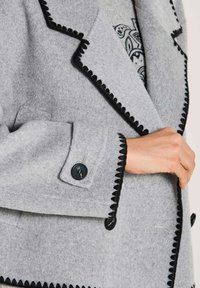 Lichtgrijze wollen jas met zwart gekarteld trim en zwarte knopen, gedragen over een gestreept shirt met zichtbaar paisley ontwerp.