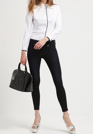 Donna che indossa una camicia bianca con bottoni e jeans skinny neri, tiene in mano una borsa nera e indossa sandali con plateau e tacco alto argentati.