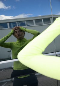 Iemand in een groen longsleeve shirt die zijn hoofd aanraakt, weerspiegeld in een glazen oppervlak buiten, met een gebouw en een blauwe lucht op de achtergrond.