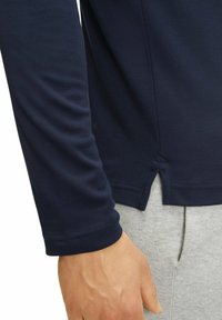 Langärmliges navyblaues Hemd mit seitlichem Schlitz; glatte Stofftextur; abgerundeter Saum; über grauen Hosen mit weichem, meliertem Finish getragen.