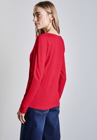 Röd långärmad tröja med rund halsringning, tillverkad av ett mjukt tyg. I kombination med blå vida jeans som visar en avslappnad passform.