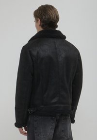 Giacca nera in finta shearling con esterno texturizzato, ampio colletto e dettaglio con fibbia in vita. Fodera morbida visibile ai polsini.