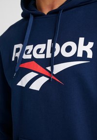 Felpa blu navy realizzata in morbido tessuto, caratterizzata da un grande logo bianco "Reebok" e un accento rosso con design grafico sulla parte anteriore, cappuccio con cordino.