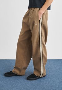 Oseba, oblečena v ohlapne rjave hlače s strančnimi črtami in napisom "obey", z rokami v žepih, stoji na modrem preprogi s črnimi čevlji.