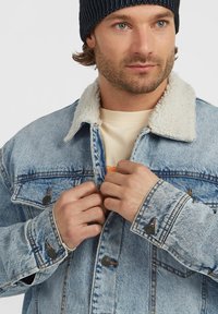 Homme avec une barbe légère attachant une veste en denim bleu clair avec un col en fourrure blanche, portant un bonnet noir et une chemise beige en dessous.