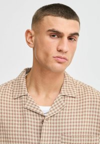 Brown and cream checkered shirt with a collar and button detail, featuring a textured fabric. Worn over a white and navy striped layer.