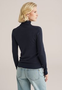 Tumši zila augsta apkakle džemperis ar šauru piegriezumu, ribotām aprocēm un apakšmalu, un mīkstu tekstūru, pārī ar gaiši ziliem džinsiem.