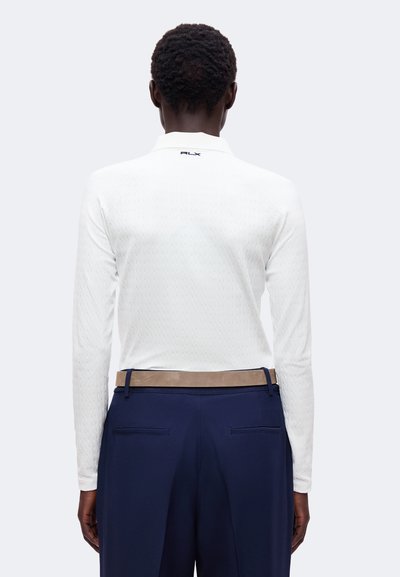 Personne portant une chemise blanche à manches longues avec le logo RLX sur le haut du dos et un pantalon bleu marine avec une ceinture beige, dos au spectateur.