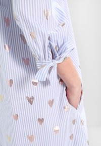 Ljusblå randig skjorta med metalliska hjärt-mönster, med knyt-detaljer vid ärmarna och en ficka på sidan.