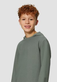 Ragazzo con capelli ricci rossi che indossa un hoodie grigio testurizzato a maniche lunghe, sorridente su uno sfondo grigio chiaro uniforme.