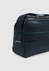 Bolso de hombro de cuero azul marino con cierre de cremallera en la parte superior, detalle de banda horizontal y adorno de metal en el lateral. Textura lisa.