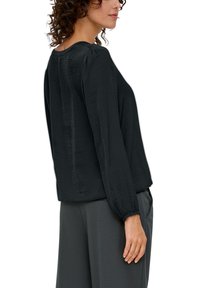 Schwarze Bluse mit langen Ärmeln, rundausschnitt und dezenter Textur, die an den Schultern geraffte Details und elastische Bündchen aufweist.