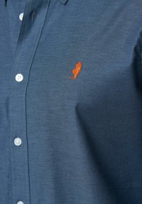 Blauwe overhemdbloes met witte knopen en een klein oranje geborduurd logo van een rechtopstaande figuur op de borst.