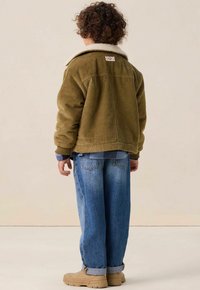 Veste en velours côtelé vert avec un col en fourrure blanche, associée à un pantalon en denim bleu et des chaussures beiges, vue de dos.