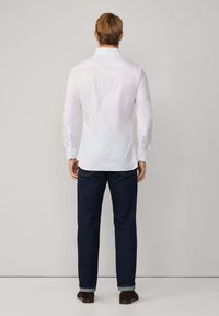 Biela košeľa s dlhým rukávom a úzkymi svetlými pruhmi, špicatý golier a gombíkové manžety, v kombinácii so sladom modrými džínsami a hnedými topánkami.