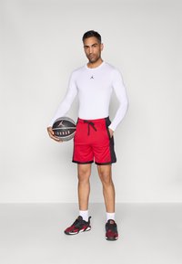 Mann i hvit langermet skjorte og røde basketballshorts holder en svart og grå Jordan basketball, stående mot en enkel bakgrunn.