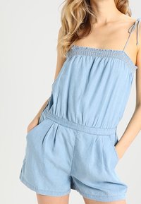 Combinaison courte en denim bleu clair avec corsage froncé, fines bretelles et poches avant à plis. Tissu décontracté et respirant.