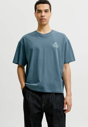 Homme portant un t-shirt bleu ample avec le texte «The Magic Fountain» et un jean foncé, debout avec une main dans la poche devant un fond uni.