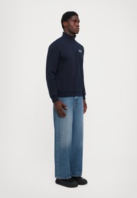 Marineblå sweatshirt med delvis glidelås i kragen, med logo på brystet. Matchet med vide, lyseblå jeans og svarte sko.
