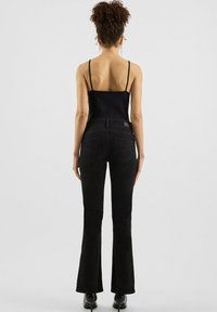 Vrouw met krullend haar, gekleed in een zwarte spaghettitop, zwarte flared jeans en zwarte hoge hakken, die met de rug naar ons toe staat.