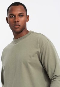 Uomo giovane con i capelli corti che indossa una camicia a maniche lunghe verde oliva, guarda dritto davanti a sé su uno sfondo bianco uniforme.