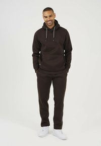 Brązowa bluza z kapturem, z przednią kieszenią i sznurkiem, w połączeniu z dopasowanymi spodniami. Nosi się z białymi sneakerami. Gładka tekstura materiału.