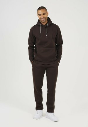 Brązowa bluza z kapturem, z przednią kieszenią i sznurkiem, w połączeniu z dopasowanymi spodniami. Nosi się z białymi sneakerami. Gładka tekstura materiału.